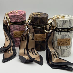 May include: Four cylindrical cases with gold-tone hardware, zippers, and attached silk scarves. The cases are pink, brown, and white with a checkered pattern. Each case has a small rectangular label that reads "Vogue New Famous."