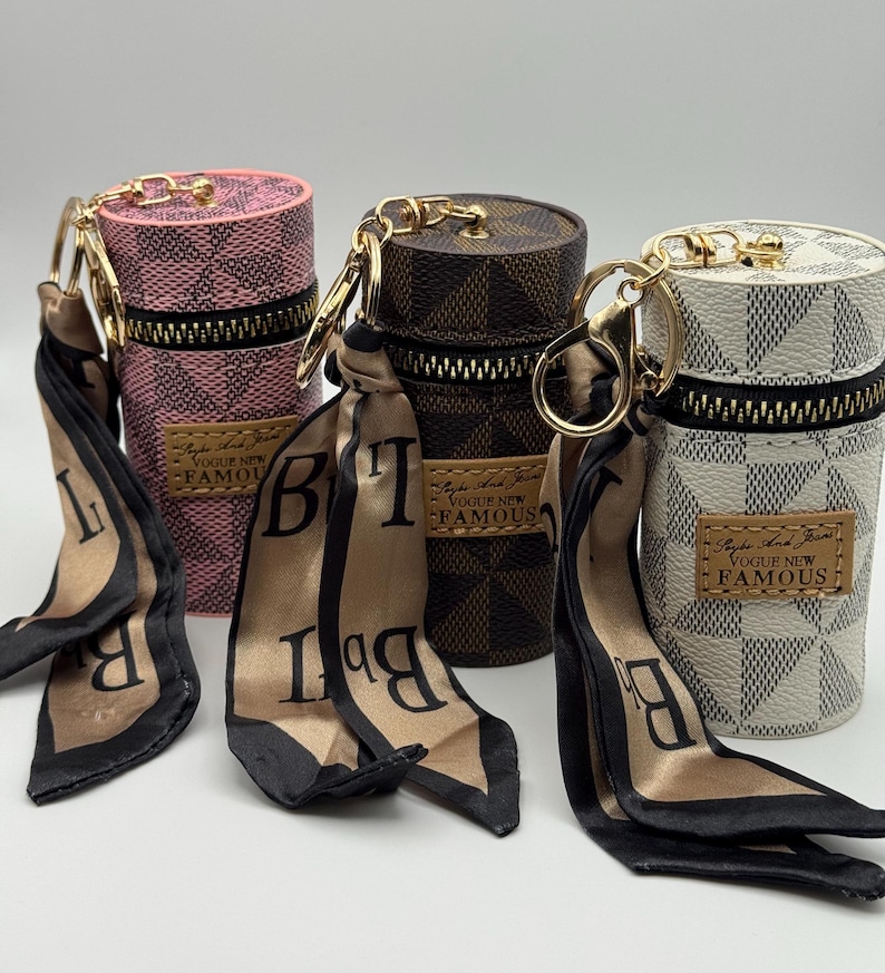 May include: Four cylindrical cases with gold-tone hardware, zippers, and attached silk scarves. The cases are pink, brown, and white with a checkered pattern. Each case has a small rectangular label that reads "Vogue New Famous."
