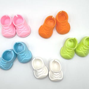 May include: A collection of colorful miniature shoes in pink, orange, green, blue, and white. The small shoes have laces and are designed for dolls or other small figures. The shoes are arranged on a white surface.