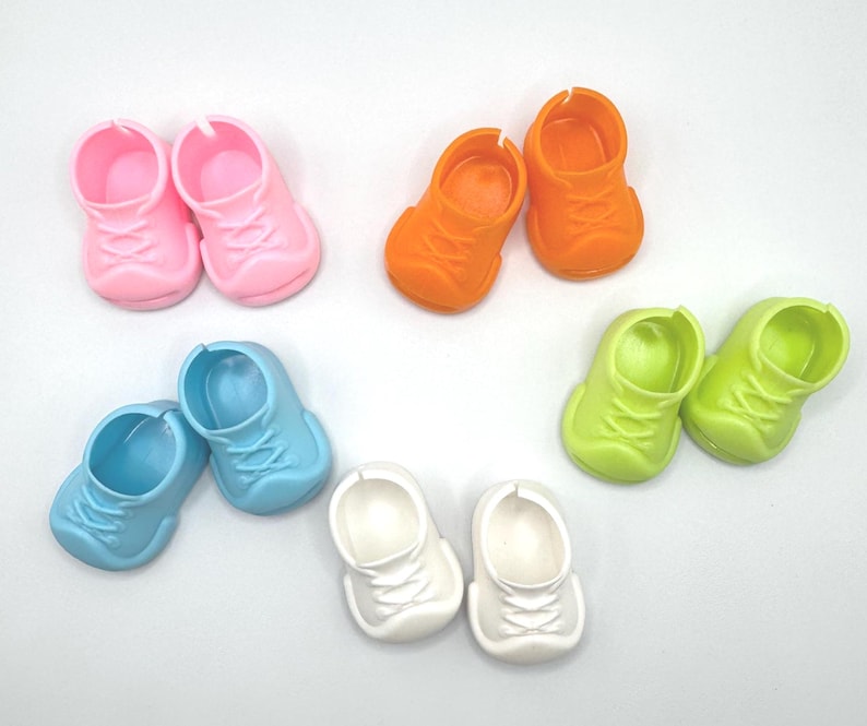 May include: A collection of colorful miniature shoes in pink, orange, green, blue, and white. The small shoes have laces and are designed for dolls or other small figures. The shoes are arranged on a white surface.