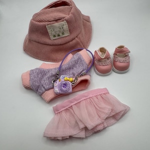 May include: A doll clothing set featuring a pink corduroy bucket hat with a bunny patch, a lavender and pink sweater, a pink tulle skirt, a purple camera purse, and pink Mary Jane shoes.