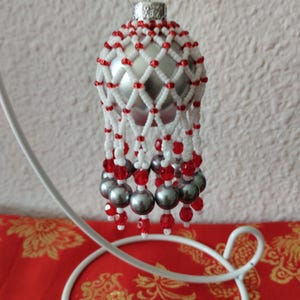 Könnte beinhalten: Silberner Weihnachtsschmuck mit einem Netzdesign aus weißen und roten Perlen. Der Schmuck hat eine silberne Basis und ist mit hängenden grauen Perlen und roten Akzenten verziert. Er wird auf einem weißen Ständer präsentiert.