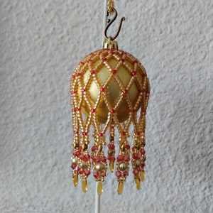 Puede incluir: Un adorno navideño dorado con un diseño de red de cuentas y detalles rojos. El adorno tiene una franja de cuentas y está colgado de un gancho dorado.