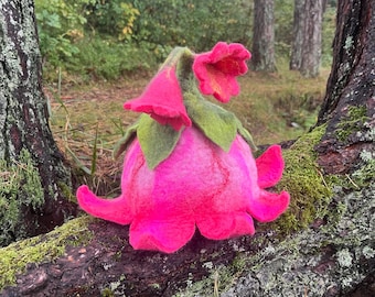 May include: A pink felt flower sculpture with green leaves, resembling a whimsical plant. The sculpture is placed on a mossy tree branch.