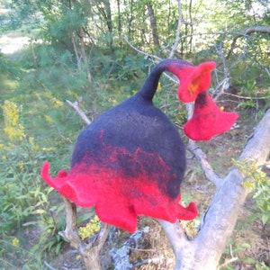 Op de afbeelding: Een zwarte en rode wollen hoed in de vorm van een bloem met een lange steel. De hoed zit op een tak in een bos.