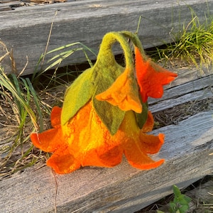 Könnte beinhalten: Eine handgefertigte Filzblumenskulptur in leuchtenden Orange-, Gelb- und Grüntönen. Die Filzblume hat mehrere Blütenblätter und Blätter und liegt auf verwitterten Holzplanken. Ein einzigartiges Kunstwerk.