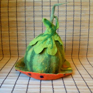 Könnte beinhalten: Ein grüner und roter Filzhut in Form einer Wassermelone mit einem grünen Filzstiel. Der Hut liegt auf einer geflochtenen Bambusmatte.