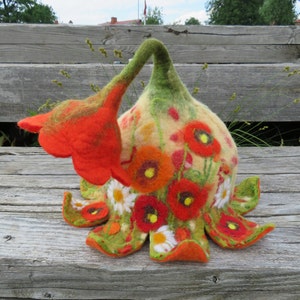 May include: A felt hat shaped like a flower with red poppies and white daisies on a cream background. The hat has a green stem and a large red poppy on the side.