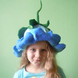 May include: A young girl wearing a blue felt flower hat with green leaves and a stem. The hat is shaped like a flower with a large, ruffled bloom. The girl has long, blonde hair and is wearing a light blue shirt.