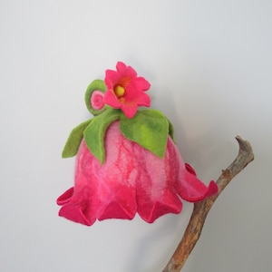 Op de afbeelding: Een roze vilten bloem met groene bladeren en een gele kern, bevestigd aan een bruine tak.