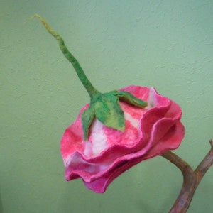 May include: A pink and white felt flower with green stem, attached to a brown branch. The flower has a ruffled edge and a white center.