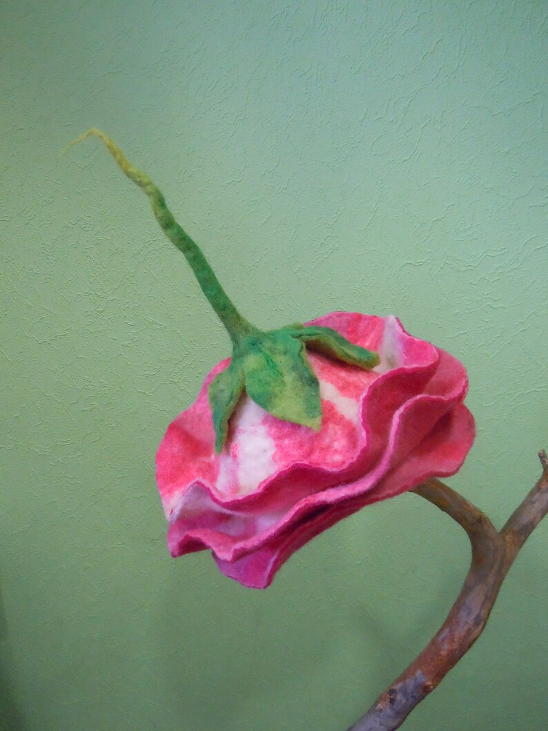 May include: A pink and white felt flower with green stem, attached to a brown branch. The flower has a ruffled edge and a white center.