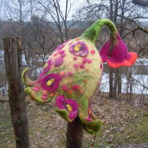 Op de afbeelding: Een fantasierijke vilten hoed in de vorm van een bloem met een groene steel en roze bloemblaadjes. De hoed is versierd met kleurrijke bloemen en stippen.