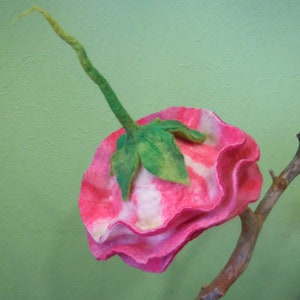 May include: A pink and white felt flower with green leaves, attached to a brown branch. The flower is made of felt and has a ruffled edge.