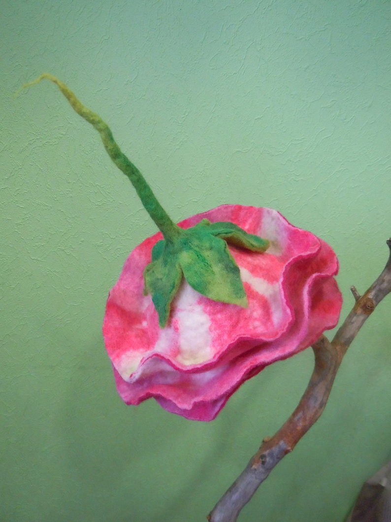 May include: A pink and white felt flower with green leaves, attached to a brown branch. The flower is made of felt and has a ruffled edge.
