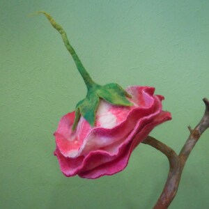 May include: A pink and white felt flower with green leaves attached to a brown branch. The flower is made of felt and has a ruffled appearance.