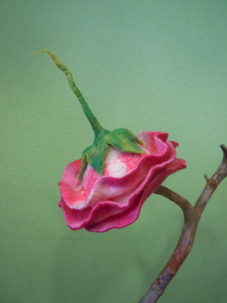 May include: A pink and white felt flower with green leaves attached to a brown branch. The flower is made of felt and has a ruffled appearance.