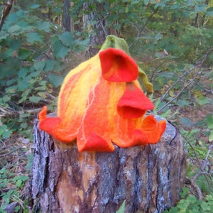 Puede incluir: Un sombrero de fieltro naranja y amarillo con forma de flor y un borde rizado. El sombrero está sentado en un tocón de árbol en un entorno boscoso.