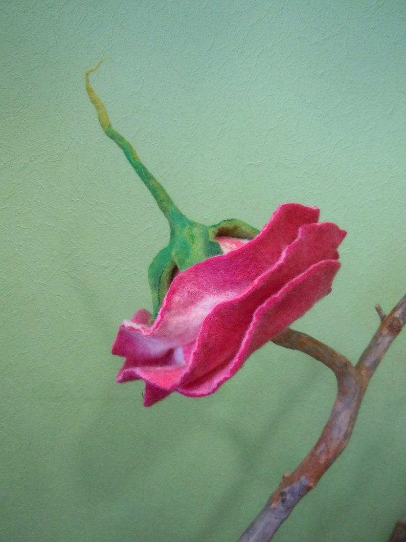 May include: A pink felt flower with green stem and leaves, attached to a brown branch.