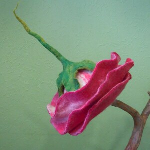 May include: A pink and white felt flower with green stem, attached to a brown branch. The flower is made of wool and has a ruffled texture.