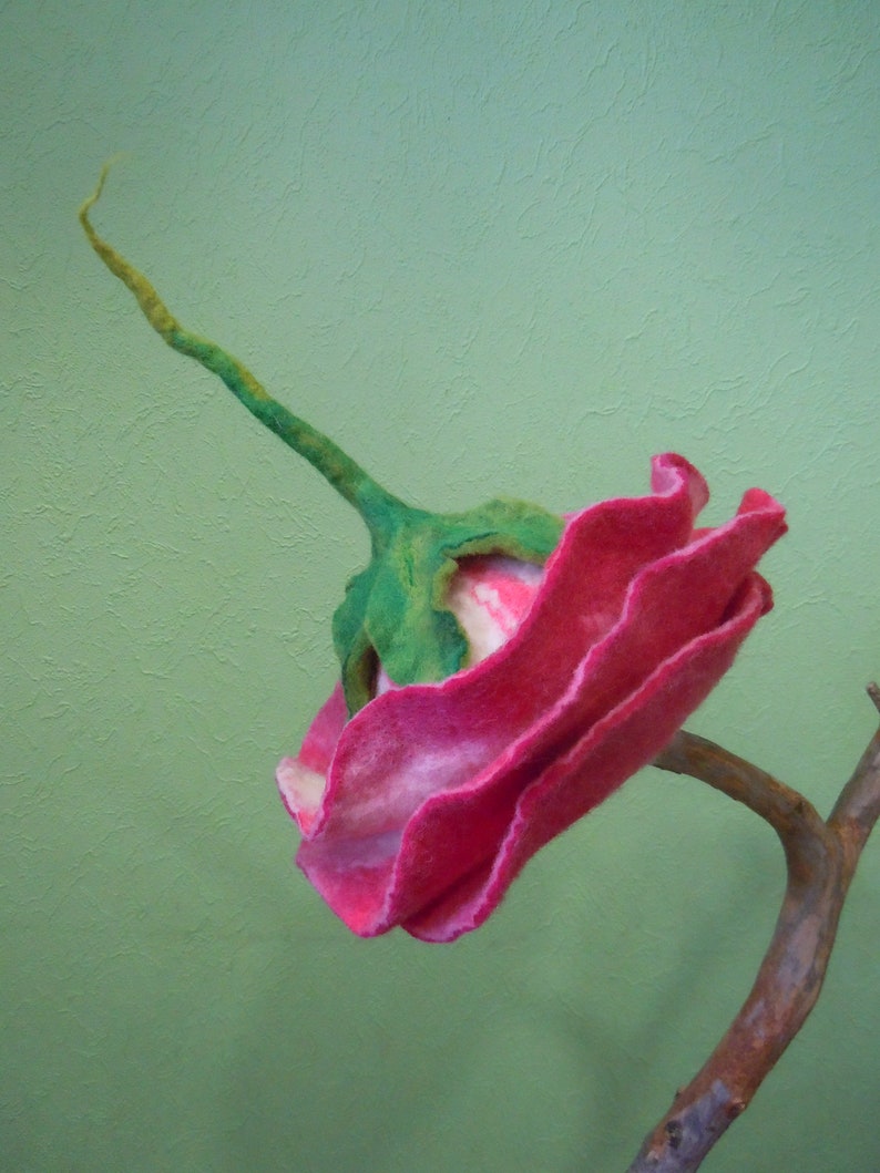 May include: A pink and white felt flower with green stem, attached to a brown branch. The flower is made of wool and has a ruffled texture.