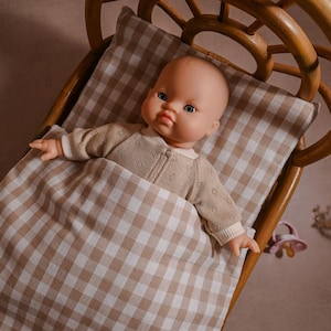 May include: A doll with brown hair and blue eyes is lying in a brown and white gingham cot. The cot is made of wicker and has a rounded shape.