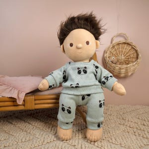 Puede incluir: Una muñeca suave que lleva un pijama azul claro con un estampado de panda. La muñeca tiene el pelo castaño y está de pie frente a una cama y una cesta de mimbre. El conjunto de pijama incluye una camiseta de manga larga y pantalones.