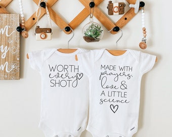 May include: Two white baby onesies with black text. The onesie on the left says "WORTH every SHOT" with a small heart. The onesie on the right says "MADE WITH prayers, love & A LITTLE science" with a small heart.