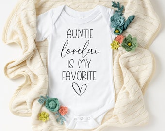 May include: White baby onesie with black text that reads "Auntie lovelai is my favorite" with a small heart below the text. The onesie is on a cream colored blanket with small green and orange felt flowers.