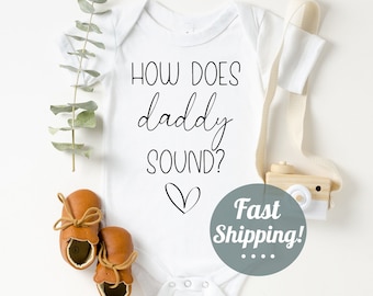 May include: White baby onesie with the text "How does daddy sound?" in black script font with a small heart underneath.  Two brown baby shoes are on the left side of the onesie.  A wooden toy camera is on the right side of the onesie.