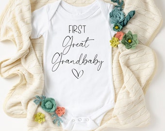 May include: White baby onesie with the text "First Great Grandbaby" in black script with a small heart underneath.