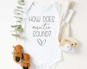 May include: White baby onesie with black text that reads "How does auntie sound?" with a small heart drawn below the text. The onesie is surrounded by brown baby shoes, a wooden toy camera, and a sprig of eucalyptus.