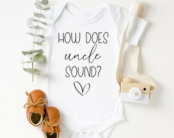 May include: White baby onesie with the text "HOW DOES uncle SOUND?" in black script font with a small heart underneath. The onesie is surrounded by brown baby shoes, a wooden toy camera, and a sprig of eucalyptus.