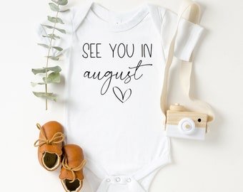 May include: White baby onesie with the text "See you in August" and a small heart. The onesie is laid flat on a white surface with brown leather baby shoes, a sprig of eucalyptus, and a wooden toy camera.
