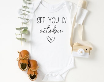 May include: White baby onesie with the text "See you in October" and a small heart. Brown leather baby shoes and a wooden toy camera are on a white background.