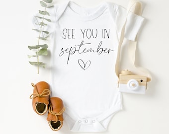 May include: White baby onesie with the text "See you in September" and a small heart.  Brown leather baby shoes and a wooden toy camera are also pictured.