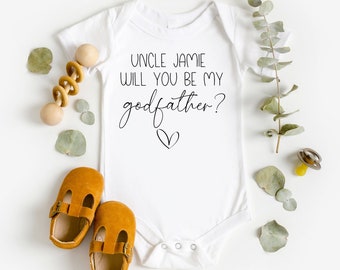 May include: White baby onesie with the text "Uncle Jamie will you be my godfather?" in black script font with a small heart below the text. The onesie is surrounded by eucalyptus leaves, a wooden rattle, and two brown baby shoes.