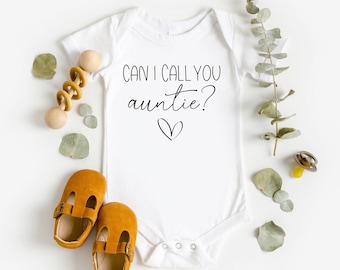 May include: White baby onesie with the text "Can I call you auntie?" in black script with a small heart underneath. The onesie is surrounded by eucalyptus leaves, a wooden rattle, and two brown baby shoes.