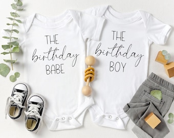 May include: Two white baby onesies with black text. The onesie on the left says "THE birthday BABE" and the onesie on the right says "THE birthday BOY".