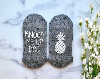 May include: A pair of gray socks with white text and a pineapple design. The text on one sock reads "Knock Me Up Doc".