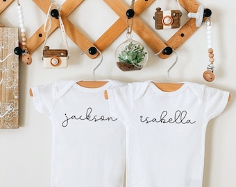 May include: Two white baby onesies with the names "Jackson" and "Isabella" written in black script. The onesies are hanging on a wooden rack with other decorative items.