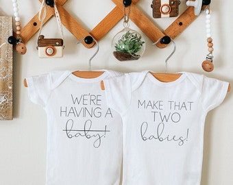 May include: Two white baby onesies hanging on a wooden rack. The onesie on the left says "We're having a baby!" with a line drawn through the word "baby". The onesie on the right says "Make that two babies!"
