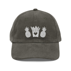 Puede incluir: Gorra de béisbol de pana verde oliva con piñas blancas bordadas y un recipiente de patatas fritas con un corazón. La gorra tiene una visera curva y una correa ajustable.