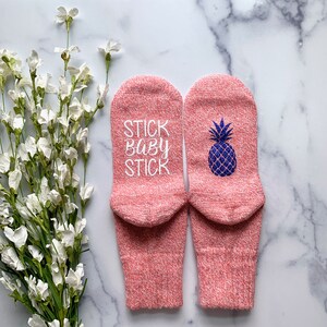 May include: A pair of pink socks with a pineapple design on one sock and the text "STICK BABY STICK" on the other sock.