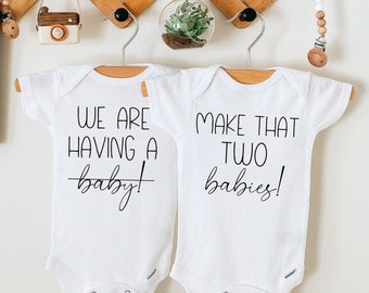 May include: Two white baby onesies hanging on hangers. The onesie on the left says "We are having a baby!" with a line through the word "baby". The onesie on the right says "Make that two babies!"