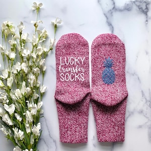 May include: A pair of pink socks with a white pineapple design on one sock and the text "LUCKY transfer SOCKS" on the other sock.