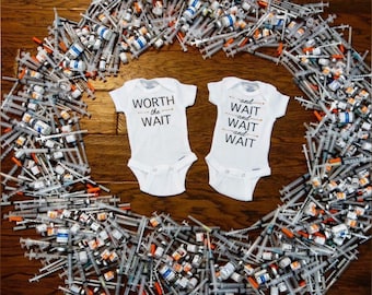 May include: Two white baby onesies with text on them. One onesie says "WORTH the WAIT" and the other says "and WAIT and WAIT and WAIT". The onesies are surrounded by a circle of syringes and vials.