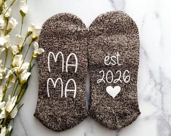 May include: Two brown and white speckled socks. One sock has the word "MAMA" embroidered in white. The other sock has "est 2026" and a white heart embroidered. The socks are on a white marble surface.