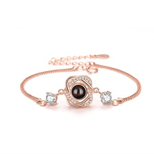 Könnte beinhalten: Ein roségoldenes Armband mit einem kleinen, runden, schwarzen Stein in der Mitte. Der Stein ist von einem zarten, klaren Kristalldesign umgeben. Das Armband hat einen Kettengliederstil und zwei kleinere, klare Kristalle auf beiden Seiten des Mittelstücks.