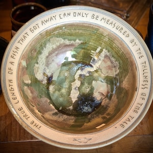 May include: A ceramic bowl with a green glaze and a white rim. The bowl has a quote inscribed on the rim: "The true length of a fish that got away can only be measured by the tallness of the tale."